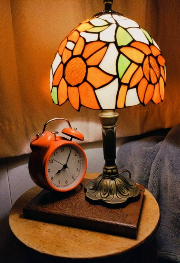 A bright red alarm clock sits beside an intricate stained-glass lamp. Many people use an alarm clock like this one to wake themselves up in the morning.
